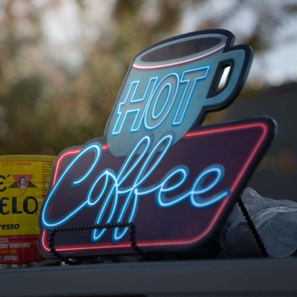 Close-up of a blue-and-red “Hot Coffee” sign shaped like a coffee cup beside a yellow coffee can at Ford of Cookeville Cars & Coffee.
