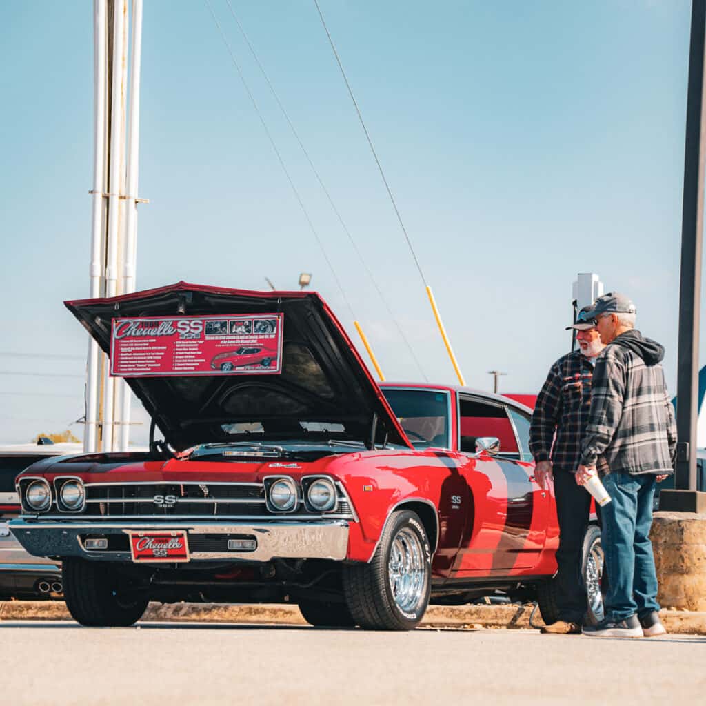 Two attendees talking beside a red 1969 Chevrolet Chevelle SS with its hood open and a vehicle information board displayed underneath at Ford of Cookeville Cars & Coffee.