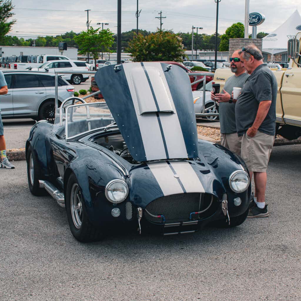 Two attendees holding drinks beside a dark blue Cobra-style roadster with silver racing stripes and its hood raised at Ford of Cookeville Cars & Coffee.