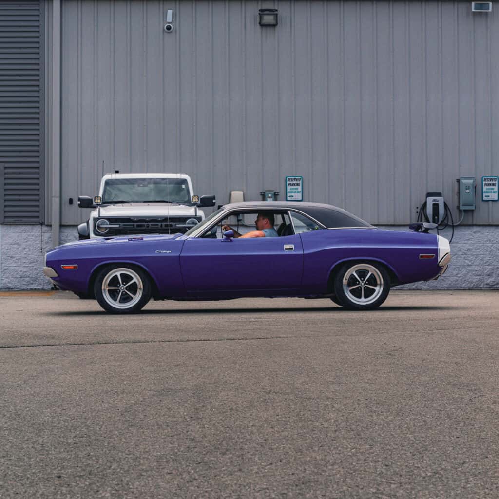 Side view of a purple classic Dodge Challenger with a black roof and a driver behind the wheel at Ford of Cookeville Cars & Coffee.