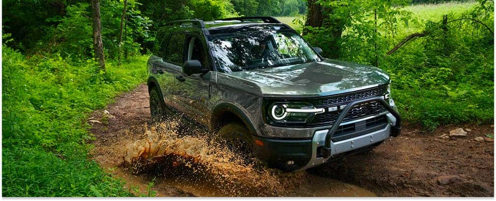 Front three-quarter view of a gray 2026 Ford Bronco Sport driving through mud on a forest trail.