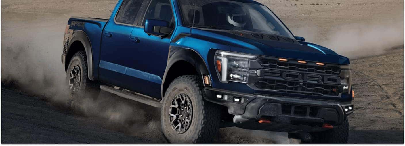 A blue 2026 Ford F-150 Raptor pickup truck with a prominent black front grille kicks up a cloud of dust while driving off-road through a desert terrain.