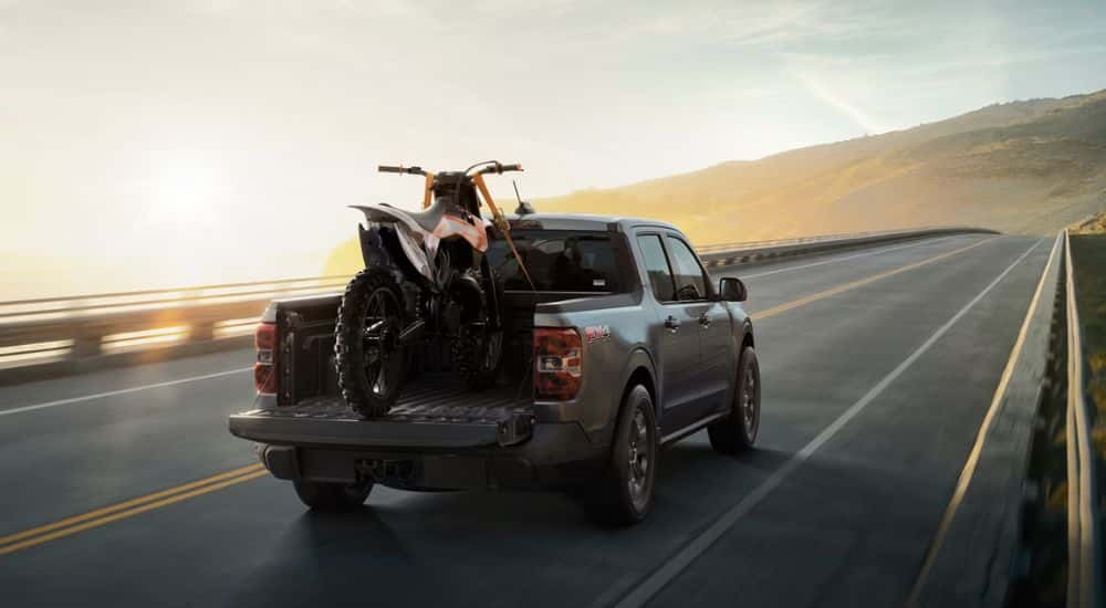 Grey 2026 Ford Maverick driving down the road with a dirt bike in the bed