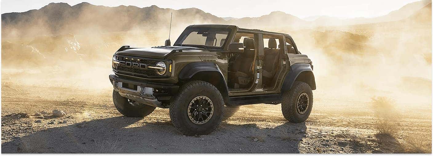 Front three-quarter view of a green 2026 Ford Bronco Raptor driving doorless on a dusty desert trail.